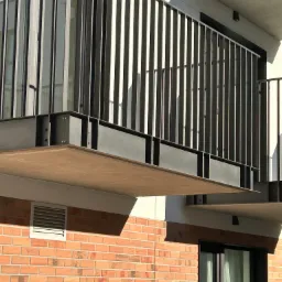 Nowoczesna, metalowa, pionowa balustrada balkonowa z elementami mocującymi, na tle ceglanej ściany i jasnego budynku. Minimalistyczny design.