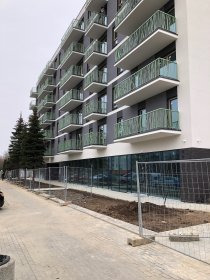 Nowoczesny budynek z balkonami z zielonymi barierkami, widok z dołu, chodnik i drzewa w tle, pochmurne niebo.