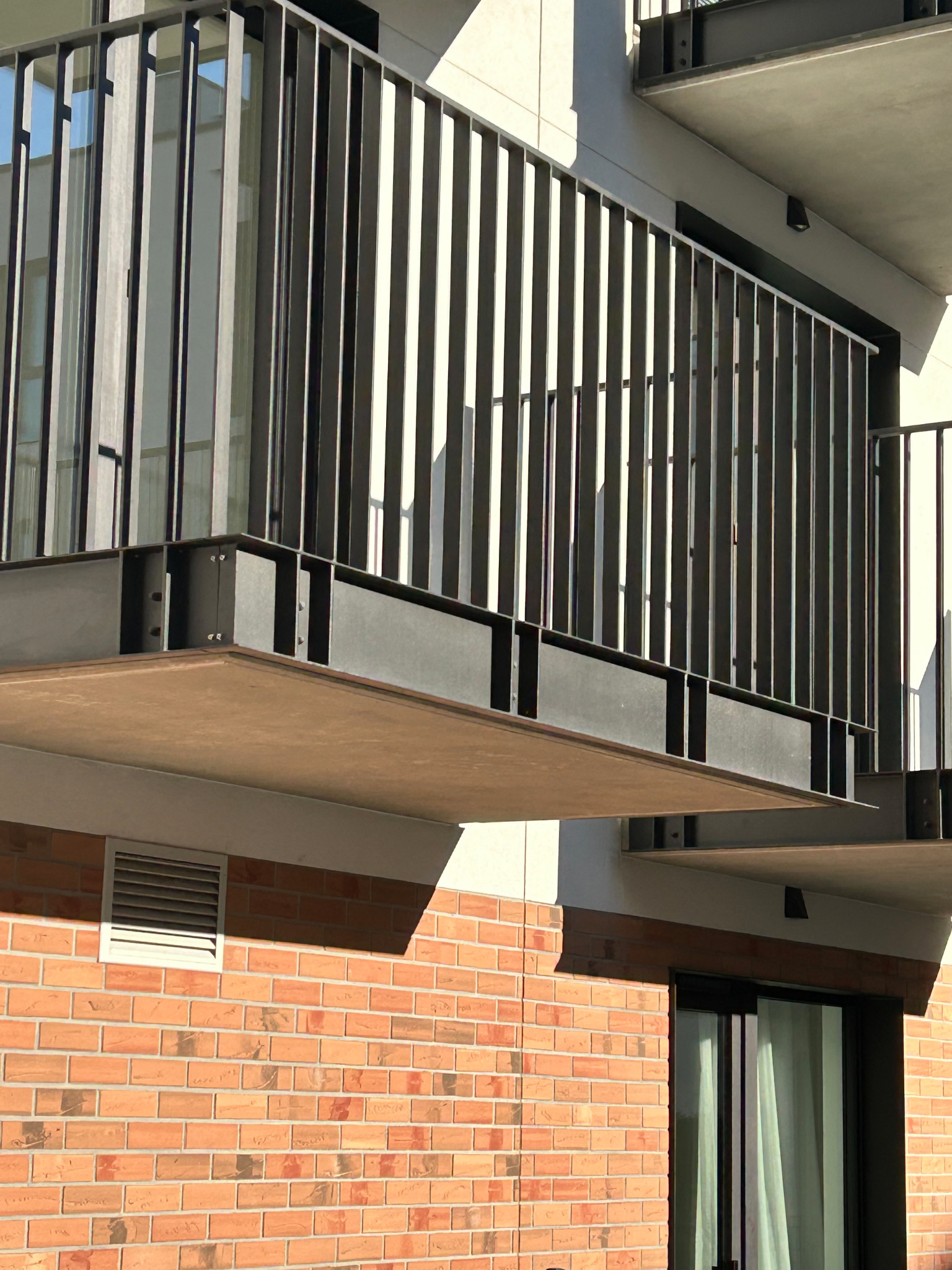 Nowoczesna, metalowa, pionowa balustrada balkonowa z elementami mocującymi, na tle ceglanej ściany i jasnego budynku. Minimalistyczny design.