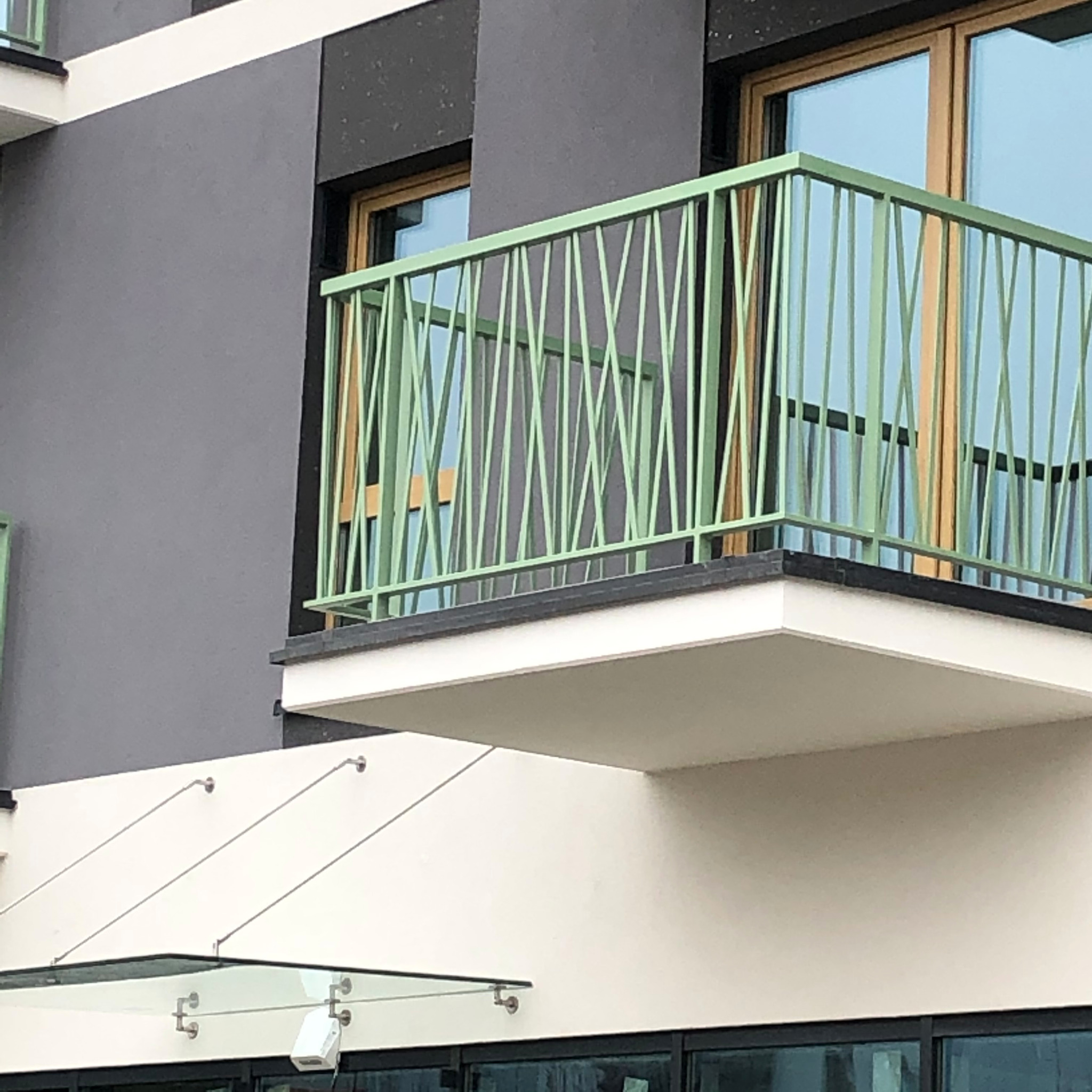 Balkon z jasnozieloną, metalową balustradą o ukośnych szczeblach, na tle szarej ściany budynku z drewnianymi oknami. Widoczny fragment szklanego zadaszenia.