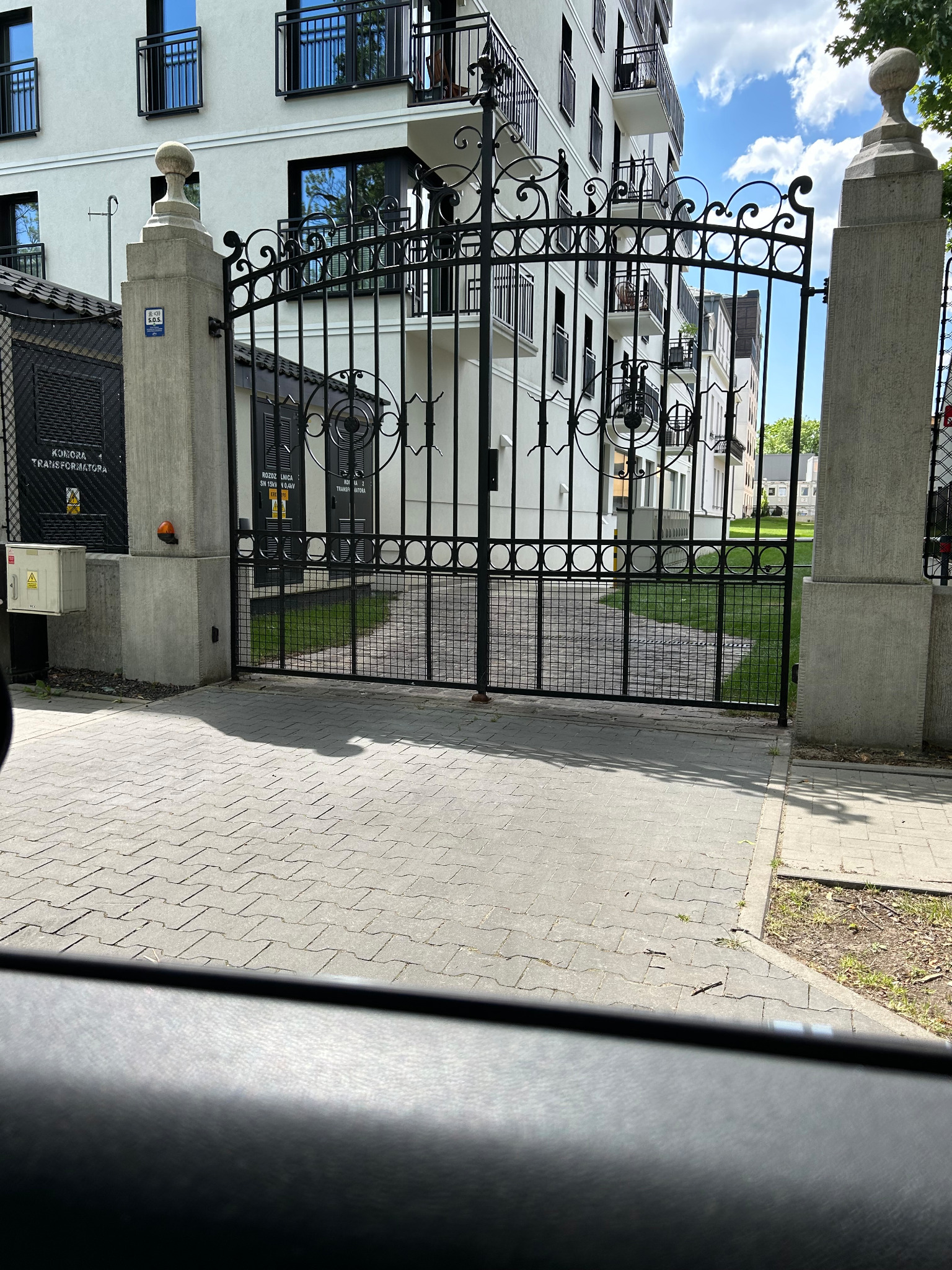 Kuta brama wjazdowa z ozdobnymi elementami, osadzona na betonowych słupach, prowadząca do nowoczesnego osiedla z białymi budynkami i balkonami.