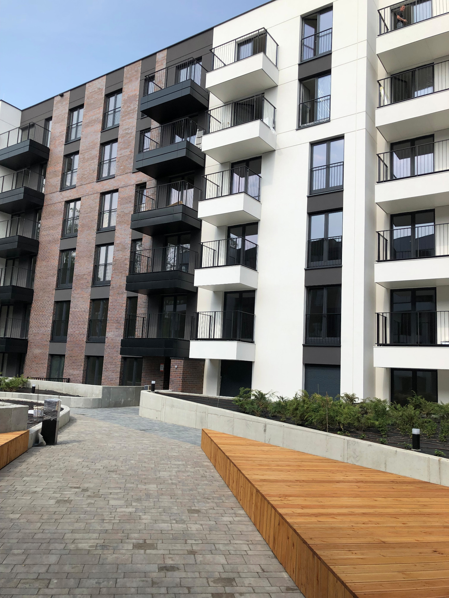 Nowoczesny budynek wielorodzinny z balkonami z czarnymi balustradami, widok z poziomu chodnika z kostki brukowej i drewnianymi elementami małej architektury.