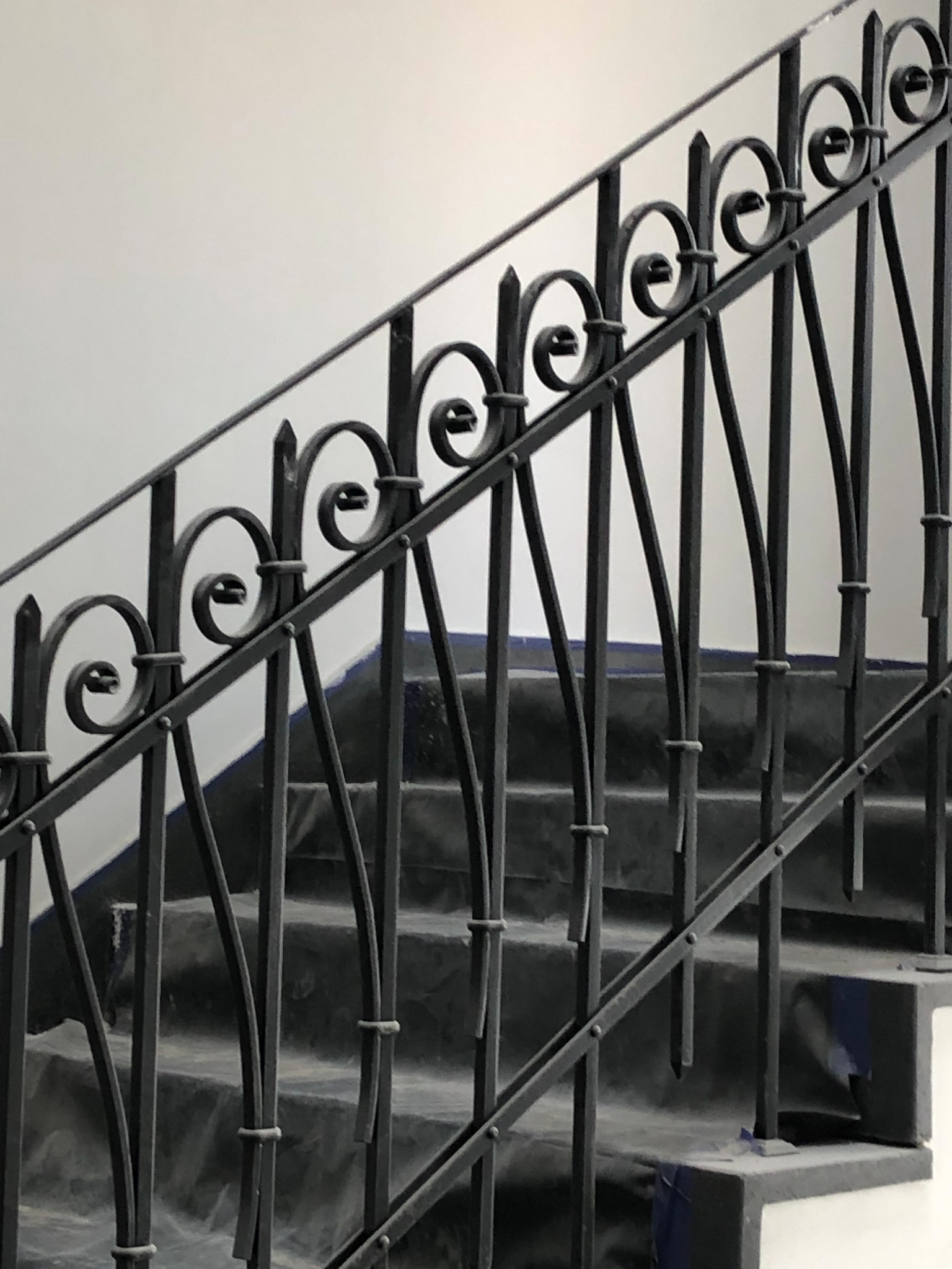 Klasyczna, czarna metalowa balustrada schodowa z dekoracyjnymi elementami w kształcie spiral, zamontowana na schodach zabezpieczonych folią malarską.