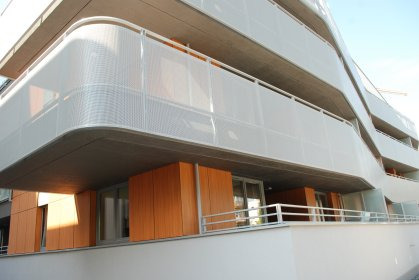 Nowoczesny budynek z balkonami osłoniętymi perforowanymi panelami, widok z dołu. Jasne, naturalne światło podkreśla architekturę.