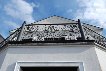 Kuta, czarna balustrada balkonowa z dekoracyjnymi elementami na tle błękitnego nieba i białej fasady budynku. Widok z dołu, podkreślający detale zdobień.