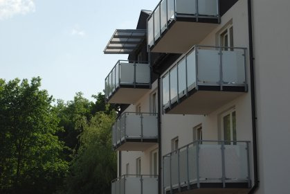 Eleganckie balkony z szklanymi panelami i metalowymi ramami, widoczne z zewnątrz budynku. Nowoczesna architektura z zielonym tłem drzew.