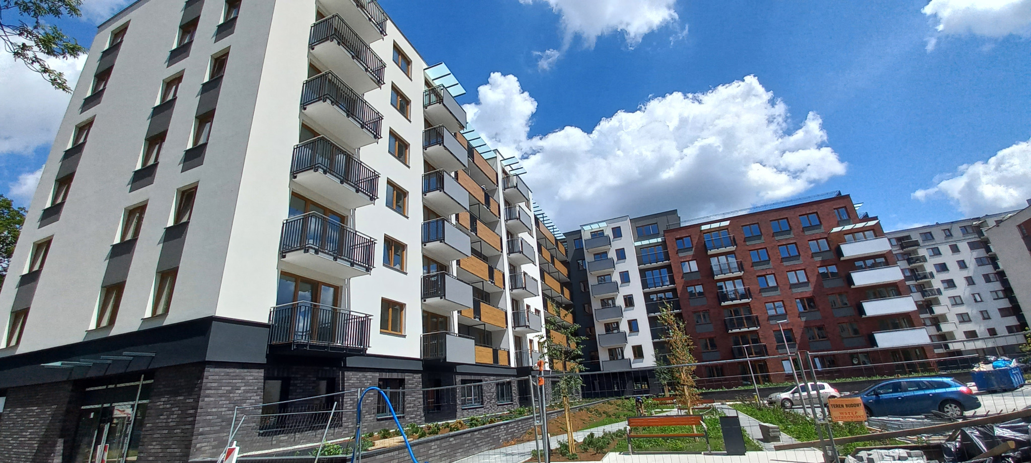 Nowoczesne budynki wielorodzinne z balkonami. Czarne, metalowe barierki balkonowe. Białe, szare i ceglane elewacje. Błękitne niebo z chmurami w tle.