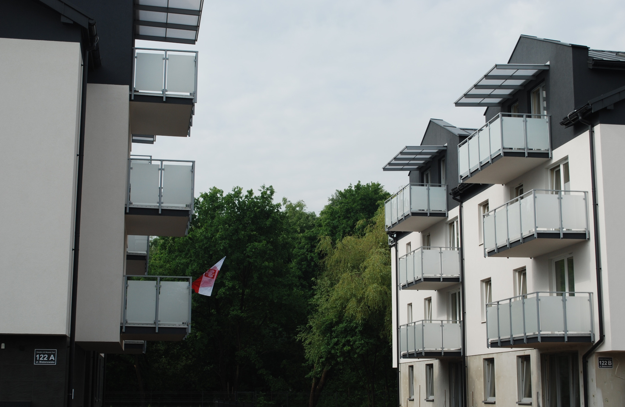 Nowoczesne balustrady szklane na balkonach apartamentowca, z widoczną flagą Polski na jednym z balkonów. Minimalistyczny design, jasna elewacja budynku.