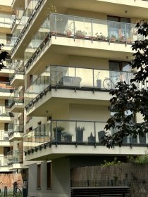 Nowoczesny budynek z balkonami, na których zamontowano minimalistyczne, szklane balustrady z czarnymi elementami mocującymi. Widok z dołu, zieleń w tle.