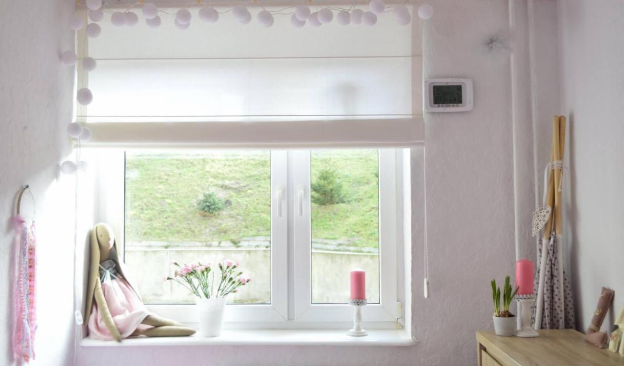 Jasny pokój dziecięcy z oknem udekorowanym girlandą z kul, na parapecie pluszowy królik, bukiet różowych kwiatów w białym wazonie i różowa świeca, za oknem zielony trawnik.