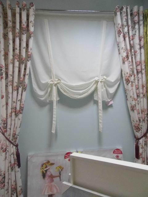 Fragment okna z dekoracyjną, podwiązywaną, białą roletą rzymską i zasłonami w kwiaty po bokach, na tle jasnoniebieskiej ściany, poniżej częściowo widoczne obrazy.