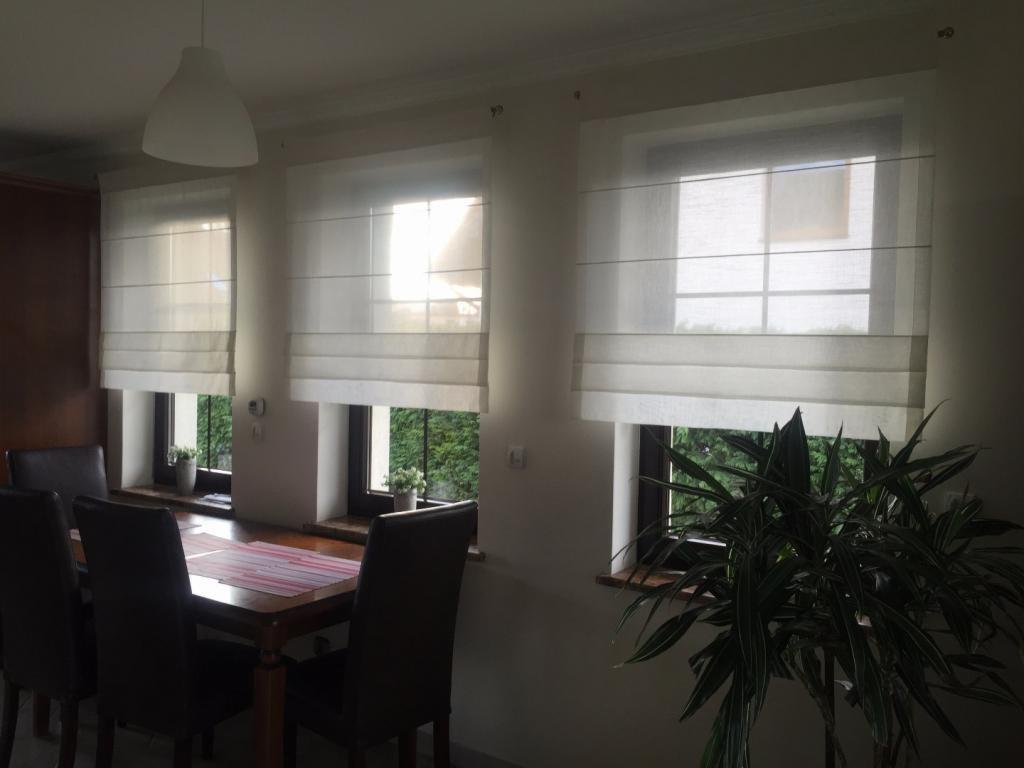 Jadalnia z trzema oknami przesłoniętymi białymi roletami rzymskimi, widok na zieleń za oknem, ciemne krzesła przy drewnianym stole, roślina doniczkowa w prawym dolnym rogu.