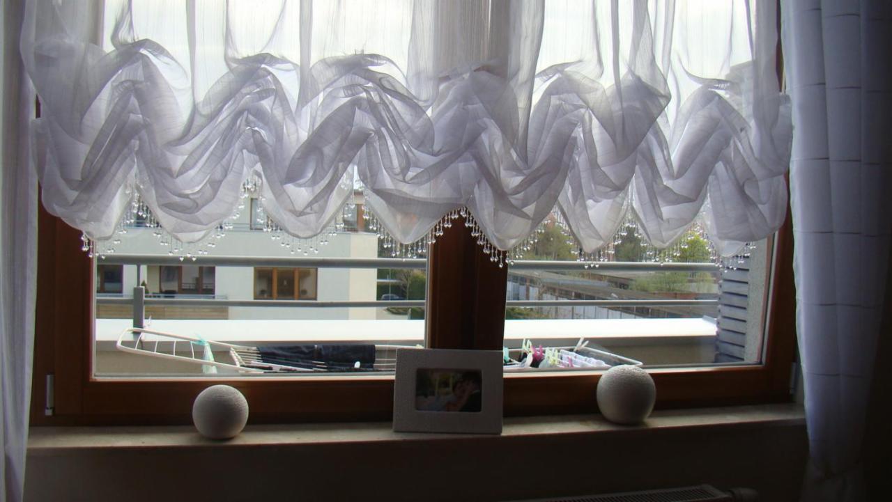 Okno z drewnianą ramą ozdobione białą, zwiewną firanką z koralikami, na parapecie dekoracyjne kule i ramka ze zdjęciem.