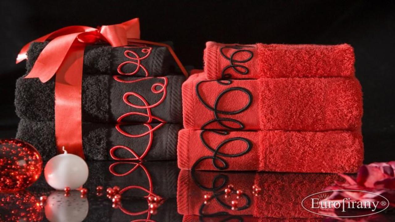 Stos ręczników w kolorach czerni i czerwieni, z dekoracyjnym haftem, przewiązanych satynową wstążką, na błyszczącej powierzchni z odbiciami świątecznych ozdób i logo firmy.