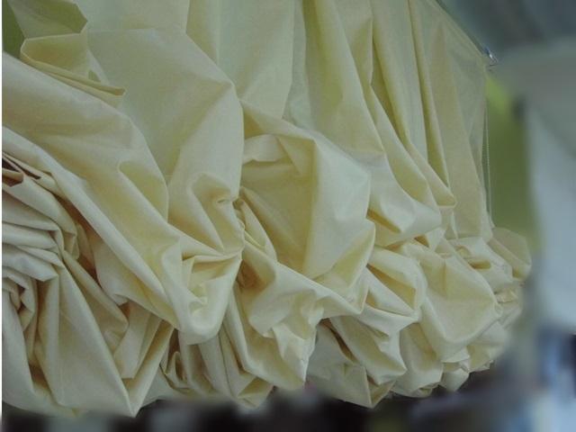 Drapowane, kremowe zasłony z błyszczącego materiału, widoczne z bliska, z delikatnym tłem w odcieniach zieleni.