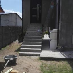 Tomasz Bud - Betonowe schody wejściowe do budynku z ciemnymi drzwiami, drabina oparta o ścianę, widoczne materiały budowlane i narzędzia.