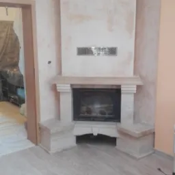 Wykończony kominek z obudową z jasnego kamienia, ściany pokryte dekoracyjnym tynkiem w odcieniach beżu, na podłodze panele, widoczne narzędzia i wiaderka po remoncie.