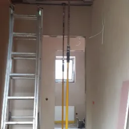 Wnętrze w trakcie montażu sufitu podwieszanego z płyt karton-gips, widoczne profile, drabina aluminiowa, podpory teleskopowe i otwór w stropie.
