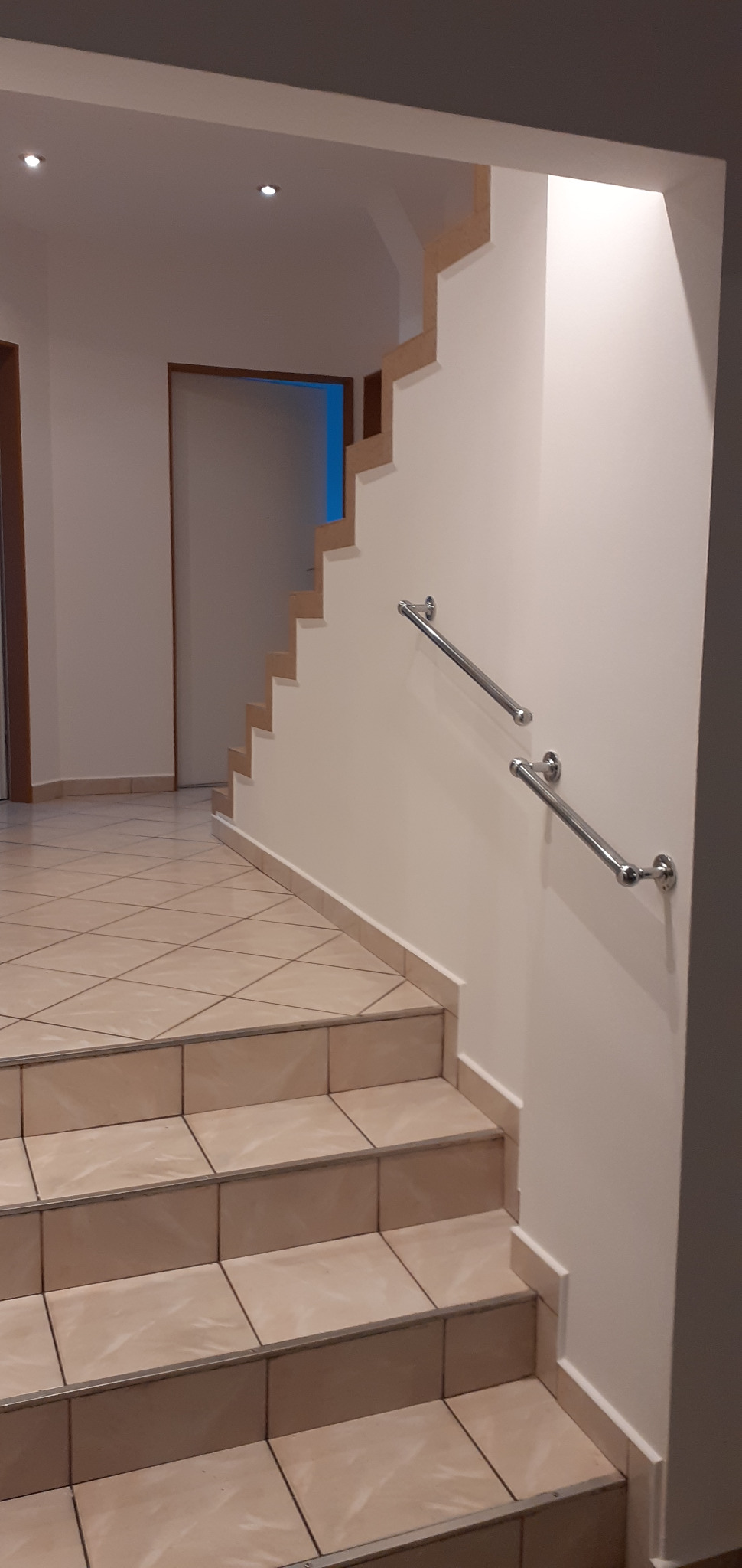 Schody wyłożone płytkami ceramicznymi w jasnym odcieniu, z metalową poręczą przymocowaną do świeżo pomalowanej ściany w jasnym kolorze.