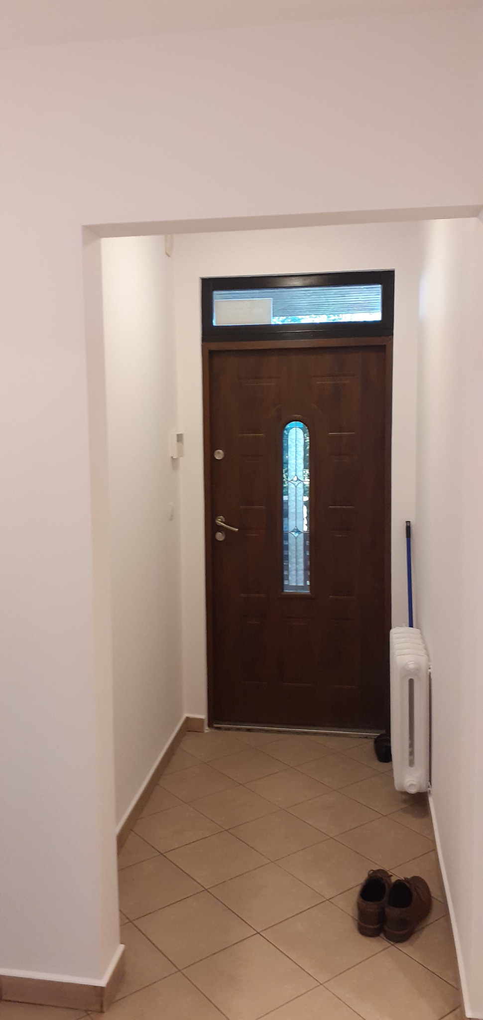 Korytarz po remoncie z pomalowanymi na biało ścianami, brązowymi drzwiami z szybą i grzejnikiem. Na podłodze leżą buty.