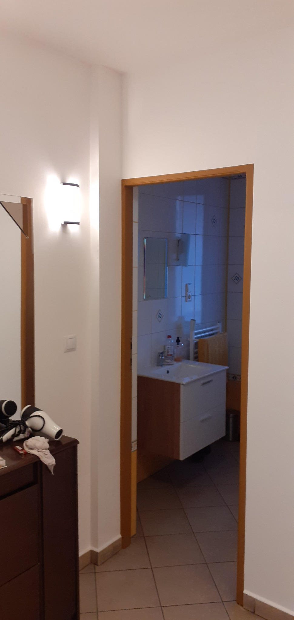 Widok na odnowioną łazienkę z umywalką w szafce, przez otwarte drzwi z drewnianą ramą. Na pierwszym planie fragment komody z suszarką do włosów.