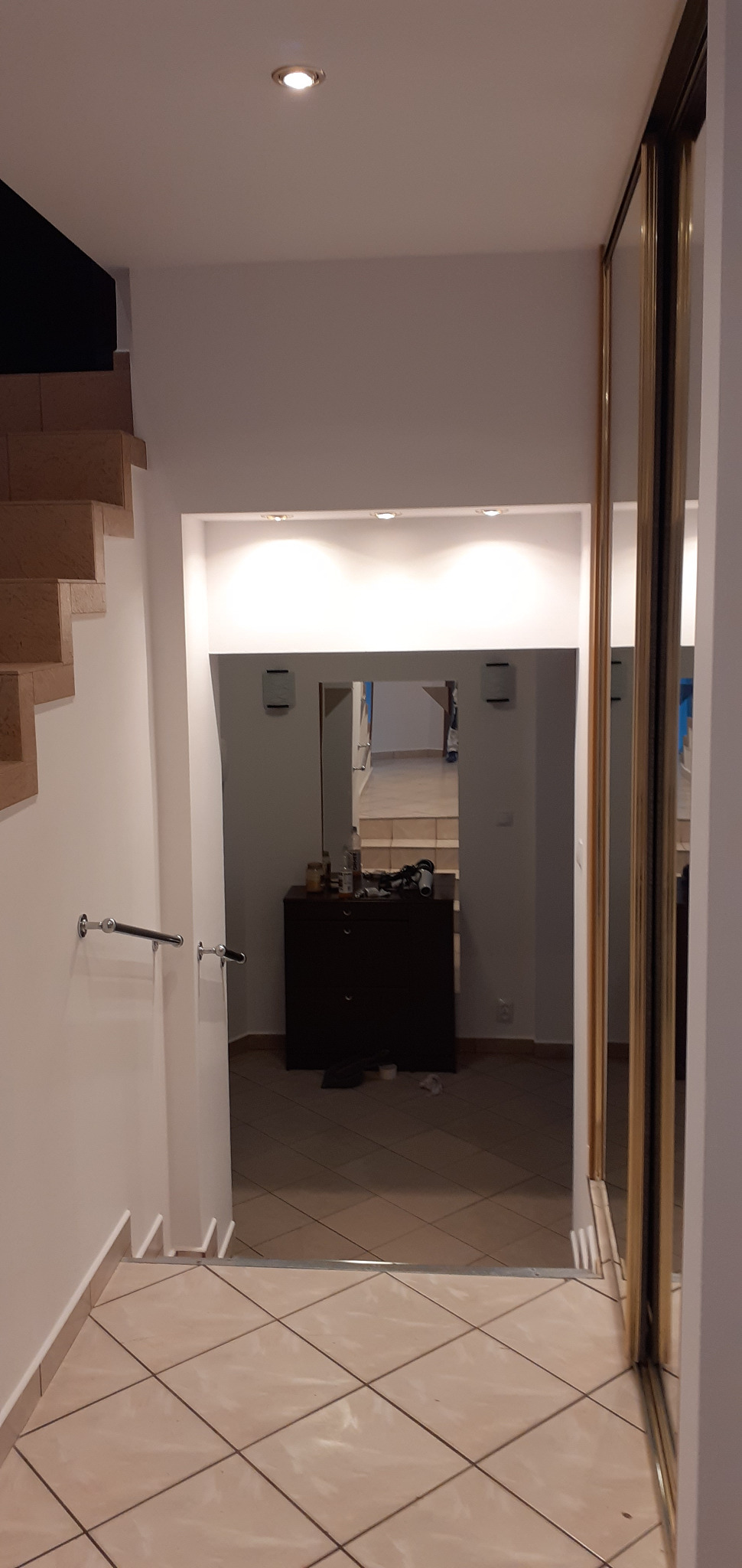 Korytarz z beżową podłogą z płytek, białe ściany z oświetleniem punktowym i lustrzana szafa przesuwna, widok na dalszą część domu z komodą.