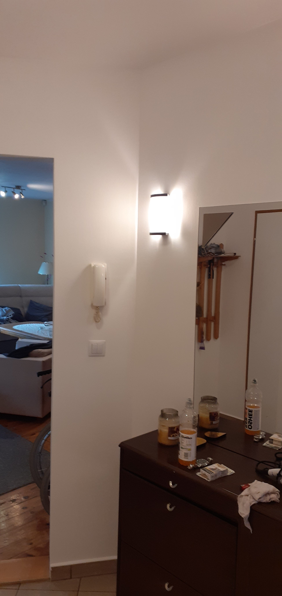 Wnętrze mieszkania: białe ściany, nowoczesna lampa ścienna, interkom, komoda z lustrem odbijającym wieszak na ubrania, widok na salon z szarą sofą.