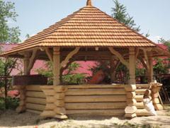 Drewniana altana ogrodowa z heksagonalnym dachem pokrytym gontem, wsparta na solidnej, poziomej konstrukcji z bali. Widoczne zdobienia w górnej części słupów. W tle zieleń drzew i czerwony dach...