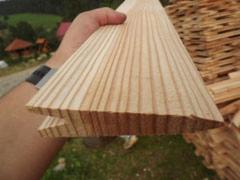 Fragment obrobionej deski z widocznym usłojeniem, trzymanej w dłoni na tle górskiego krajobrazu i stosu drewna.