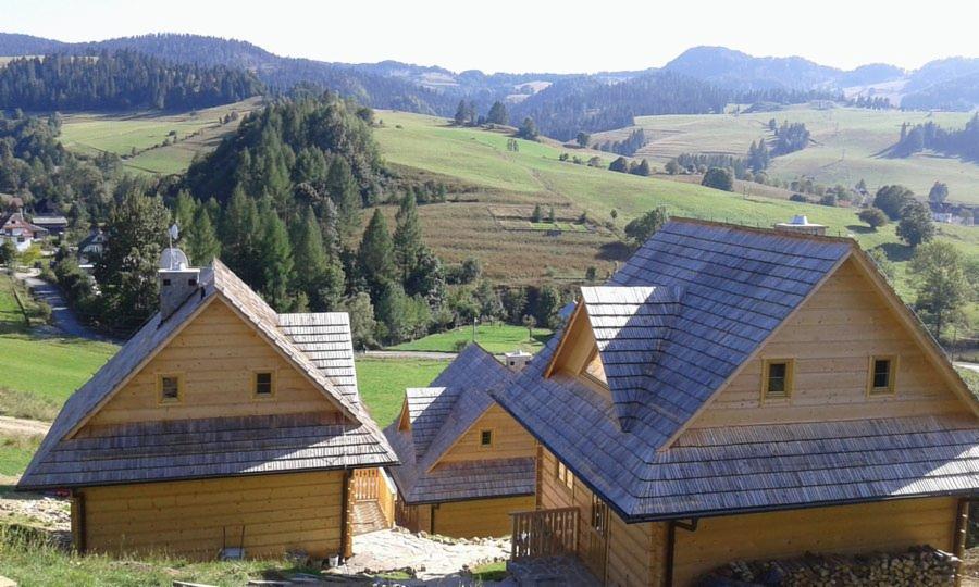 Szereg drewnianych domków w stylu góralskim z szarymi dachami gontowymi, na tle zielonych wzgórz i lasów.