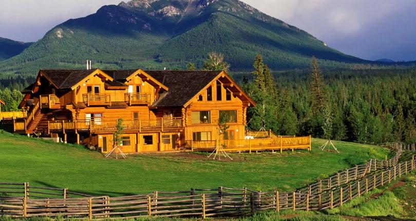 Dwupiętrowy dom z bali z ciemnym dachem i balkonami, otoczony zielonym trawnikiem i drewnianym płotem, na tle zalesionych gór.