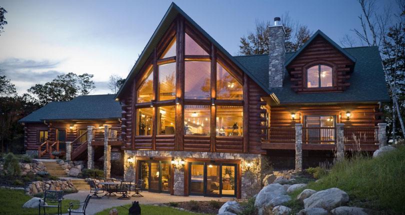 Dwupiętrowy dom z bali z dużymi oknami i kamienną podmurówką, oświetlony wieczornym światłem, otoczony zielenią i skałami.