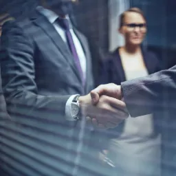 Uścisk dłoni dwóch mężczyzn w garniturach, widoczny przez żaluzje, z kobietą w tle, symbolizujący zawarcie umowy lub partnerstwo biznesowe.