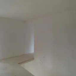 Surowe wnętrze w trakcie prac wykończeniowych: białe ściany po nałożeniu gładzi, widoczne przewody elektryczne, okno z widokiem na zewnątrz, surowa podłoga przed położeniem posadzki.