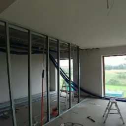 Wnętrze w trakcie budowy z widoczną metalową konstrukcją ścian działowych, niebieskie rury wentylacyjne pod sufitem, surowe ściany, okna z widokiem na zewnątrz i rozstawione drabiny.