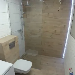 Nowoczesna łazienka z kabiną prysznicową typu walk-in, wykończona płytkami imitującymi drewno, z widoczną toaletą i częściowo zamontowanym oświetleniem LED na ścianie.