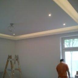 Primo-Rem - Nowoczesny sufit podwieszany z oświetleniem LED, widoczna drabina malarska i fragment sylwetki osoby w pomarańczowej koszulce przy oknie.