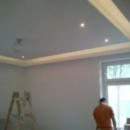 Nowoczesny sufit podwieszany z oświetleniem LED, widoczna drabina malarska i fragment sylwetki osoby w pomarańczowej koszulce przy oknie.