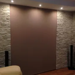 Ściana w salonie z dekoracyjną okładziną kamienną po bokach i gładką, brązową powierzchnią pośrodku, oświetlona punktowymi lampami sufitowymi, z fragmentem beżowej sofy w rogu.