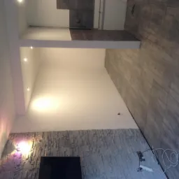 Wykończone wnętrze pokoju z szarą kamienną okładziną na ścianie, szarą podłogą z płytek imitujących kamień oraz białymi ścianami i sufitem z punktowym oświetleniem