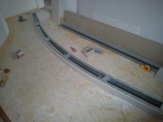 Przygotowanie do montażu profili sufitu napinanego o nietypowym, łukowatym kształcie. Widoczne profile aluminiowe, narzędzia i płyty gipsowo-kartonowe w surowym wnętrzu.