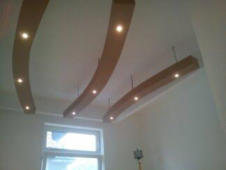 Biały sufit z dekoracyjnymi, podwieszanymi elementami z drewna w kształcie łuków, oświetlonych punktowymi lampami LED, widok z dołu na tle okna.