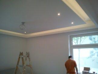 Nowoczesny sufit podwieszany z oświetleniem LED, widoczna drabina malarska i fragment sylwetki osoby w pomarańczowej koszulce przy oknie.