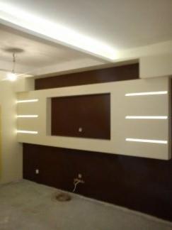 Nowoczesna ściana z podświetlanym panelem dekoracyjnym w kolorach brązu i beżu, widoczne gniazdko elektryczne na ścianie, sufit z oświetleniem LED.