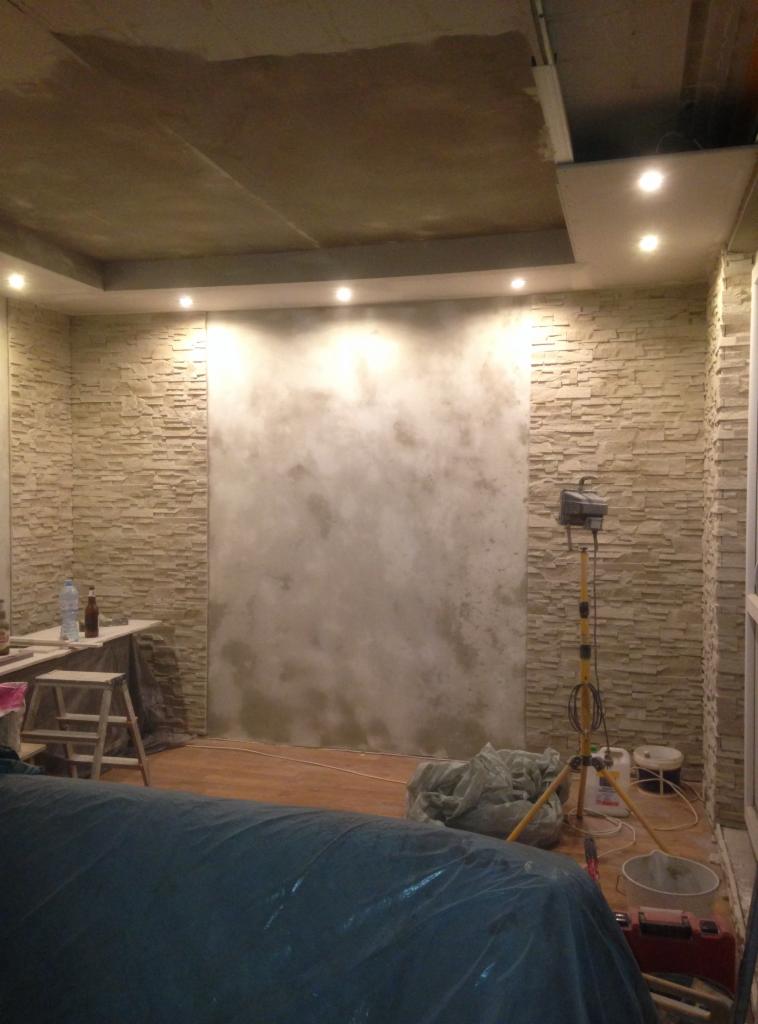 Wnętrze w trakcie remontu: ściany częściowo wykończone kamieniem dekoracyjnym, fragment ściany z nałożoną gładzią, widoczne narzędzia i oświetlenie robocze.