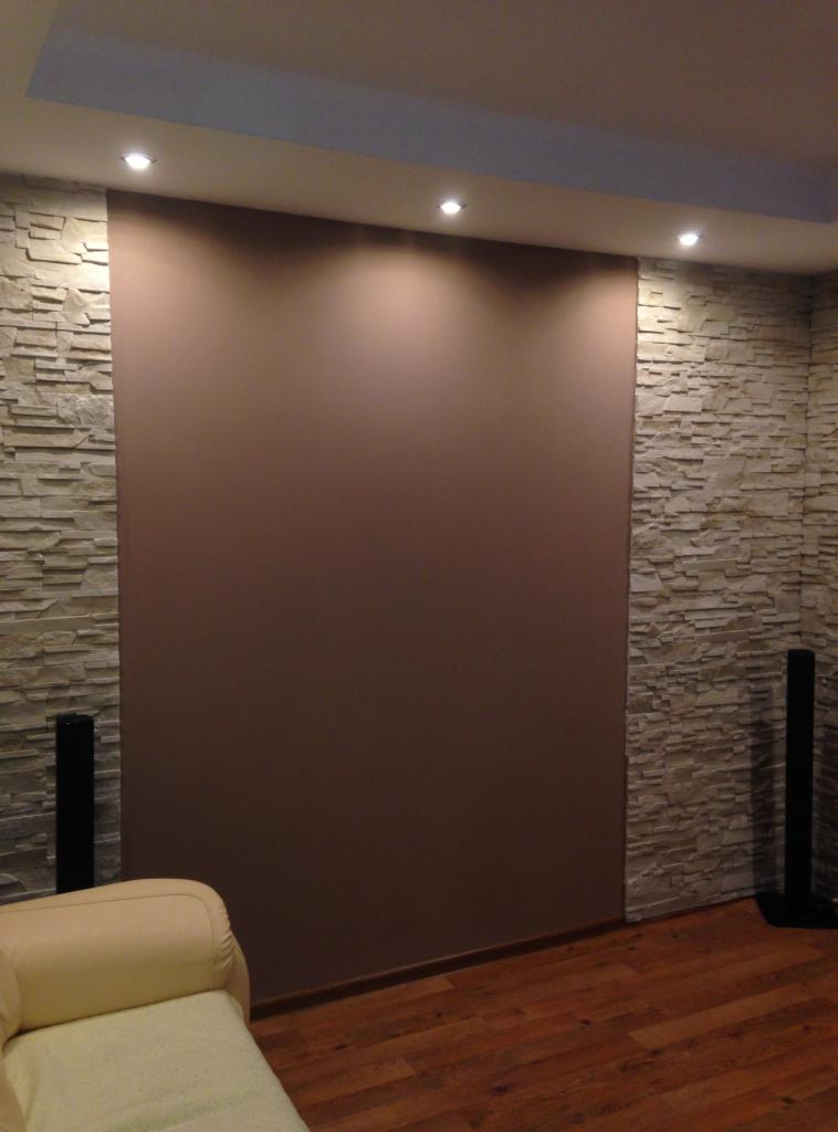 Ściana w salonie z dekoracyjną okładziną kamienną po bokach i gładką, brązową powierzchnią pośrodku, oświetlona punktowymi lampami sufitowymi, z fragmentem beżowej sofy w rogu.