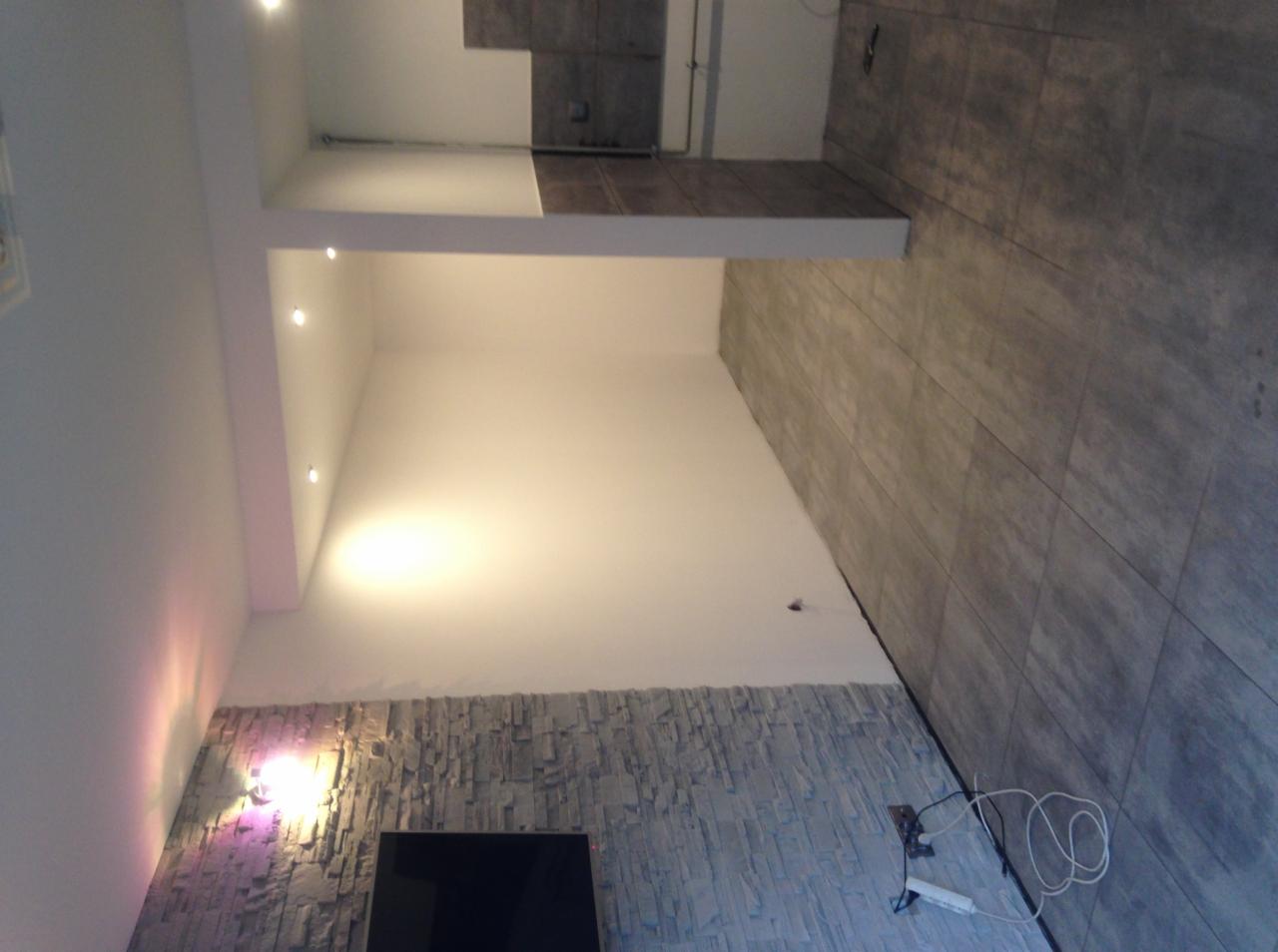 Wykończone wnętrze pokoju z szarą kamienną okładziną na ścianie, szarą podłogą z płytek imitujących kamień oraz białymi ścianami i sufitem z punktowym oświetleniem