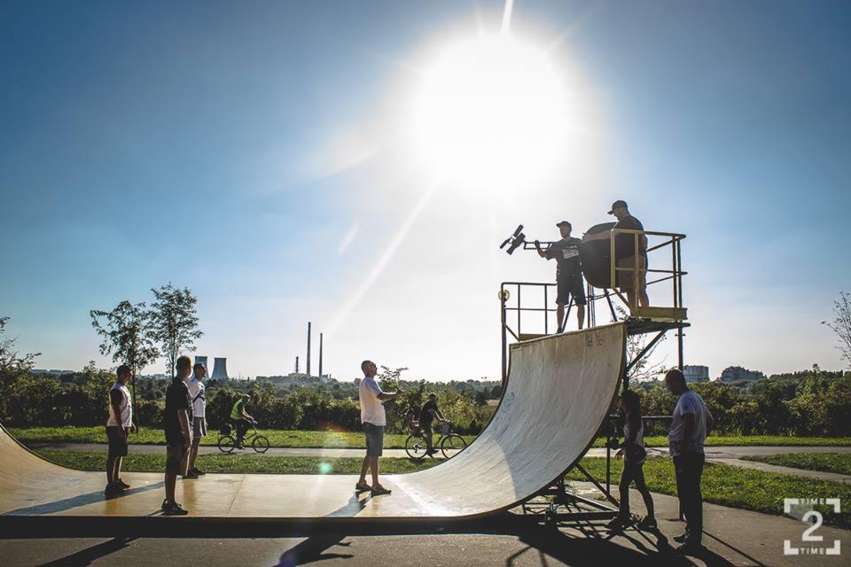 Ekipa filmowa kręci ujęcia na rampie skateparku; operator z kamerą i asystent z blendą na platformie, grupa osób obserwuje, w tle kominy elektrociepłowni.