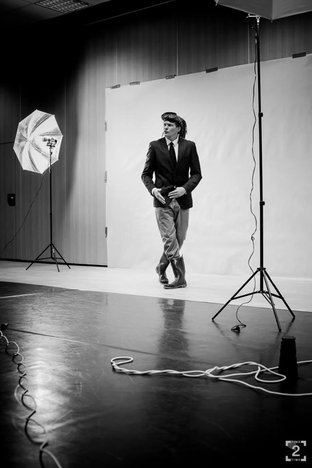 Czarno-białe zdjęcie mężczyzny w garniturze i kaloszach pozującego w studiu fotograficznym z lampą błyskową i parasolką.