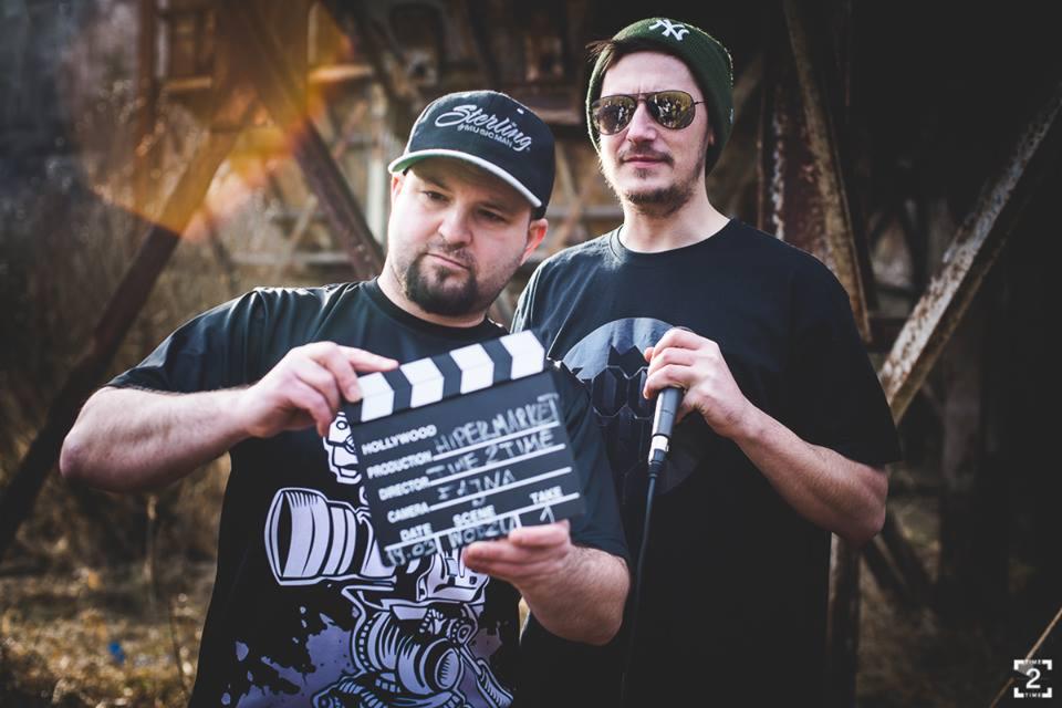 Dwóch mężczyzn na planie filmowym: jeden trzyma klaps filmowy z napisem 'Hipermarket 2 Time', drugi z mikrofonem, w tle metalowa konstrukcja